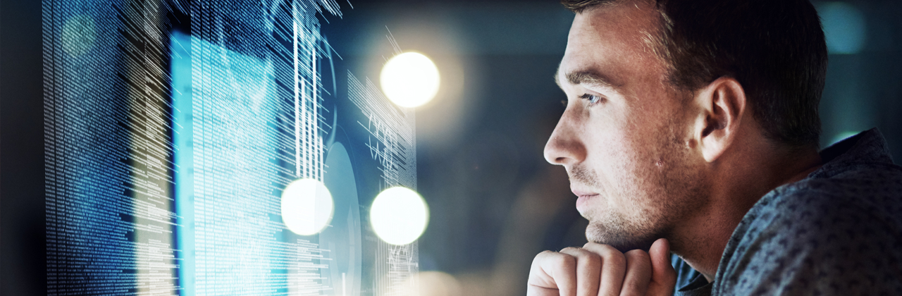 A person sits at a desk closely studying lines of code displayed on a large monitor in a dimly lit workspace.