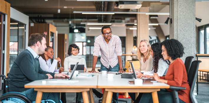 Bright open concept office. There is a work meeting being held where a group of diverse people, including a man on a wheelchair are working together.