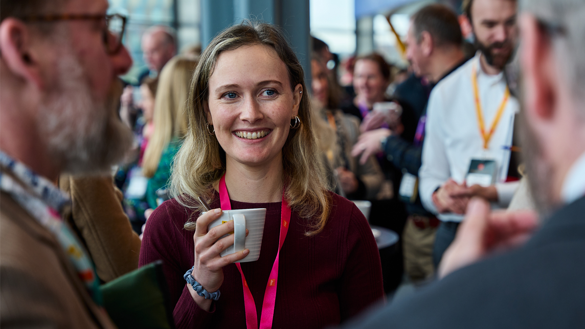 Person holding a white mug at a busy indoor event, wearing a lanyard, with several people conversing in the background.