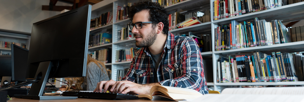 A man stares intently at a computer screen surrounded by books in a library.