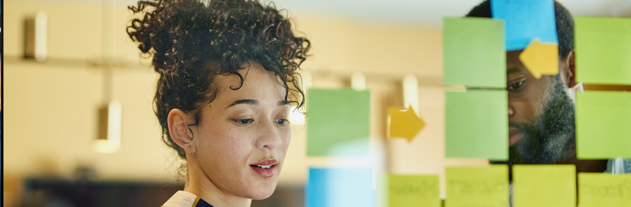 A man and a woman are standing in front of a glass wall writing on sticky notes.