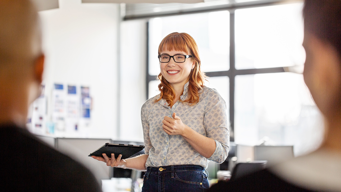 A woman with a tablet addresses an audience of work colleagues.